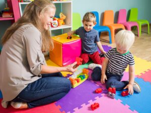 https://doradcy365.pl/wp-content/uploads/2022/04/two-little-boys-are-playing-with-kindergarten-teacher-woman-is-sitting-with-boys-floor-playroom-min-scaled-300x225.jpg